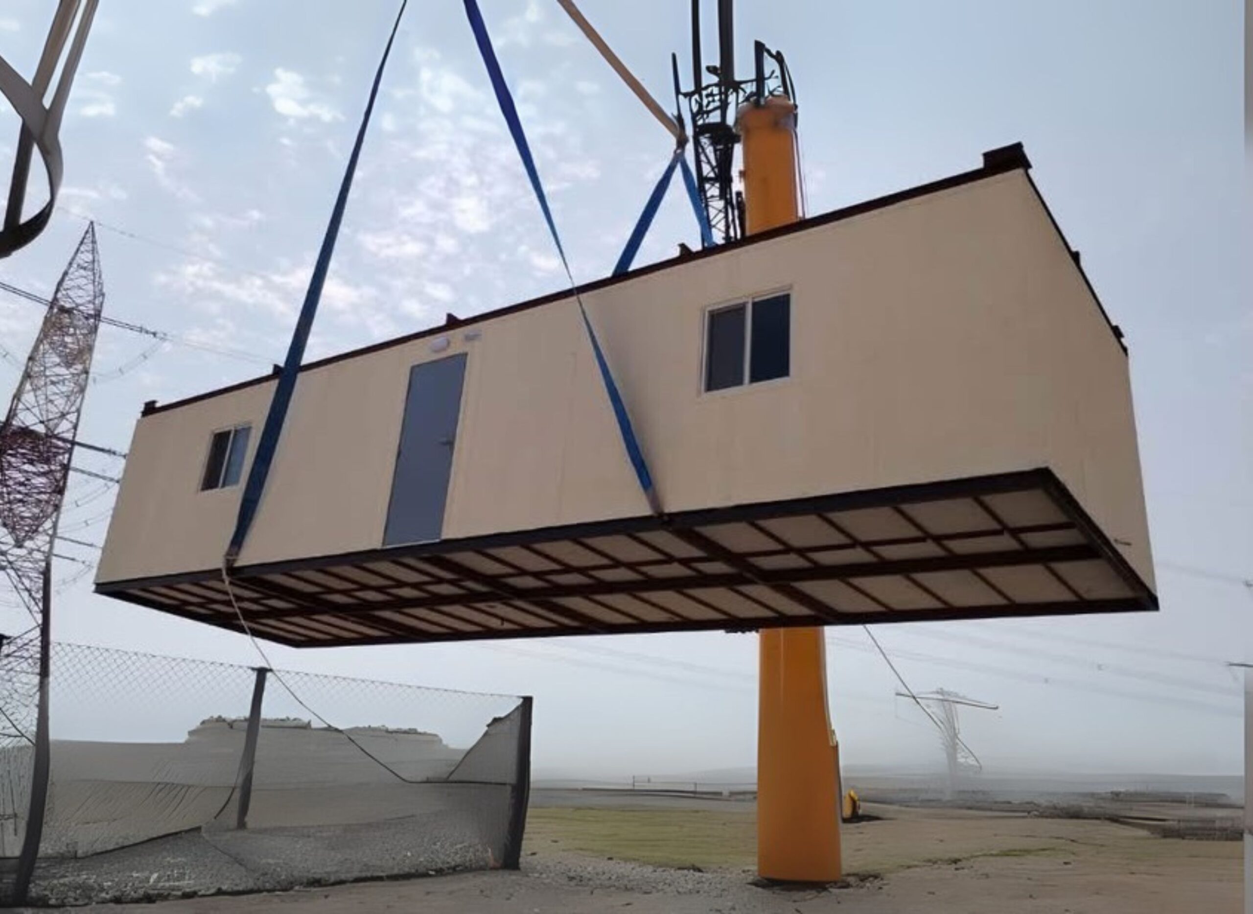 A construction unit is suspended from a crane, with a clear sky in the background and power lines visible in the distance.
